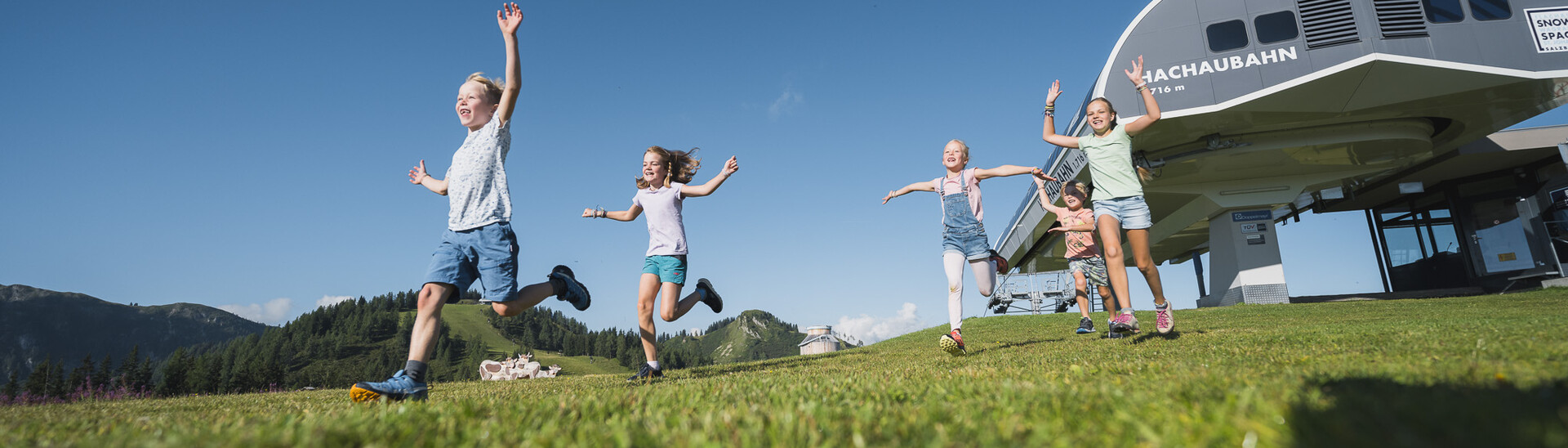 Vier Kinder springen vor Freude über die Wiese, hinter ihnen die Blackaubahn im Snow Space Salzburg bei strahlendem Sonnenschein