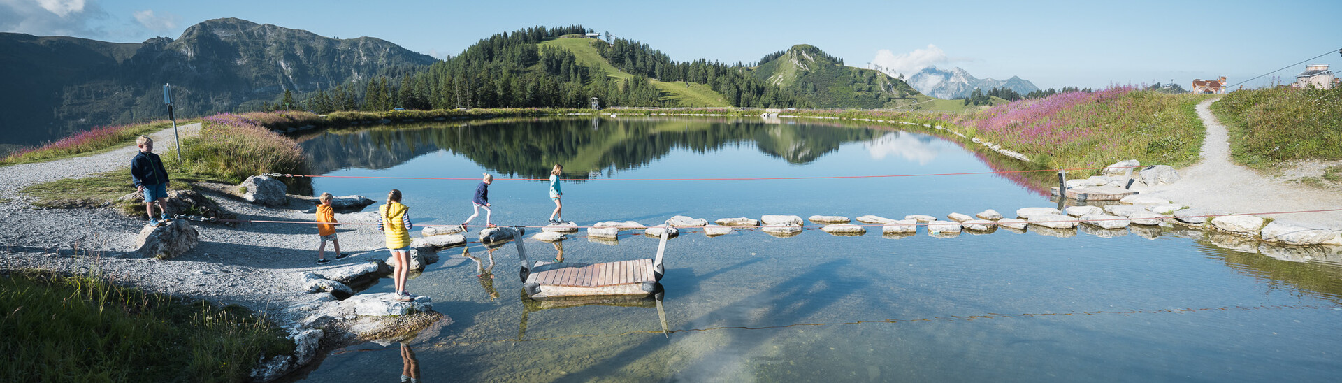 Besucher überqueren auf Baumstämmen einen stillen Bergsee im Snow Space Salzburg, umgeben von einer malerischen Berglandschaft